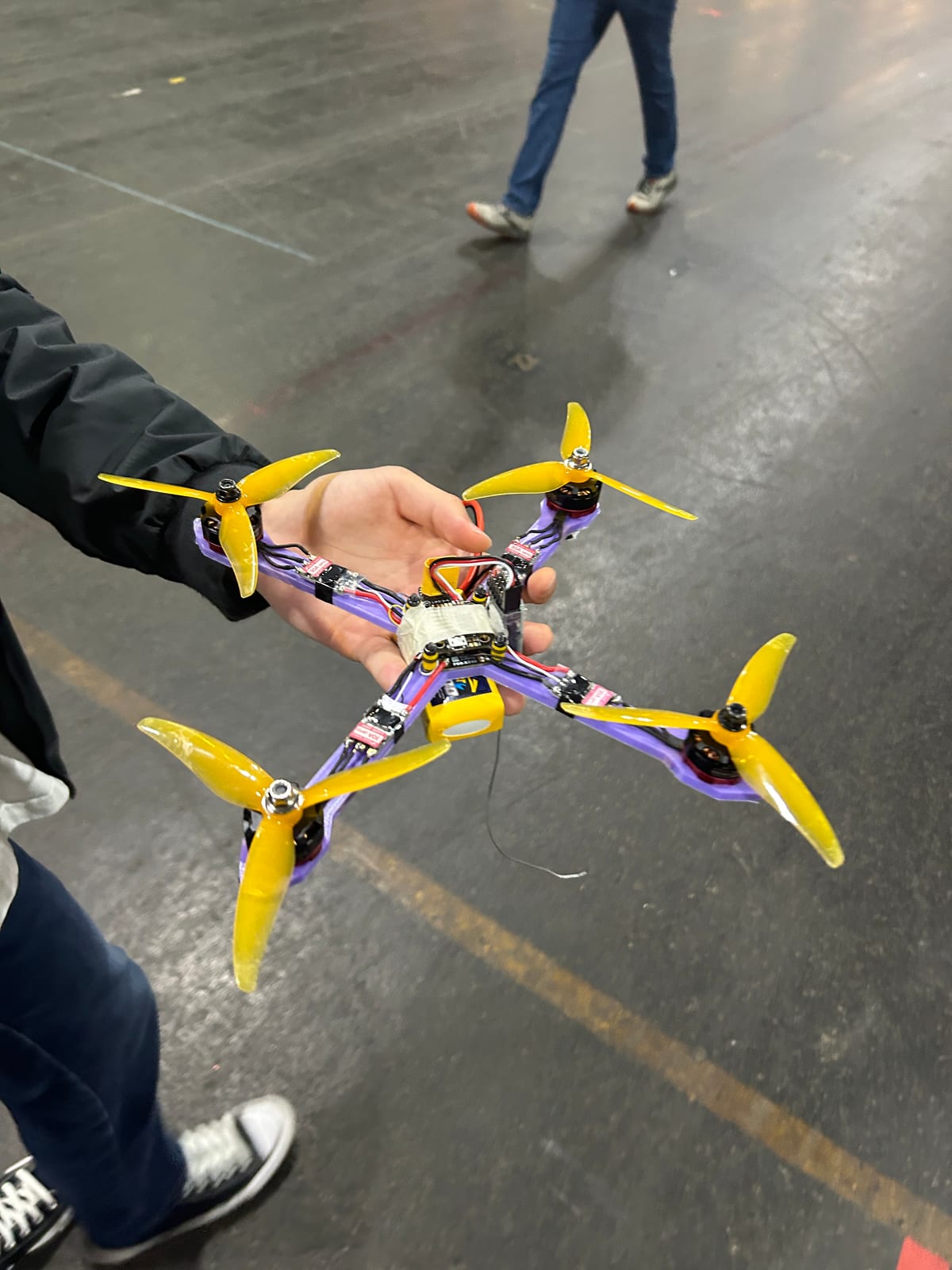 A quadcopter with a purple 3D-printed frame and yellow three-blade propellers held up in one hand on a competition floor, motors and wiring visible along the arms, with people and a taped course line on the concrete below.