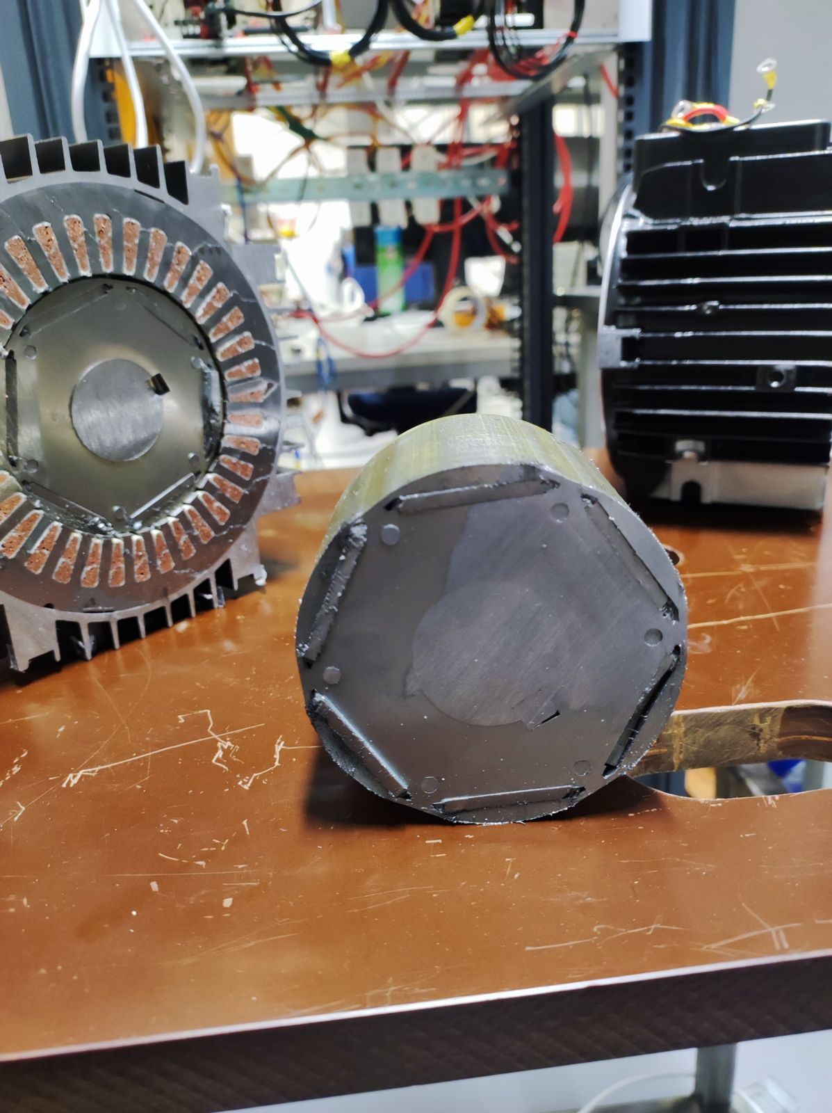 Photograph of the physical IPMSM rotor sectioned across its axis, resting on a workbench in front of the machine's sectioned stator and its finned black housing. The cut face shows the near-hexagonal rotor lamination stack with the buried permanent-magnet pockets and their retaining bridges visible as dark slots around the perimeter.
