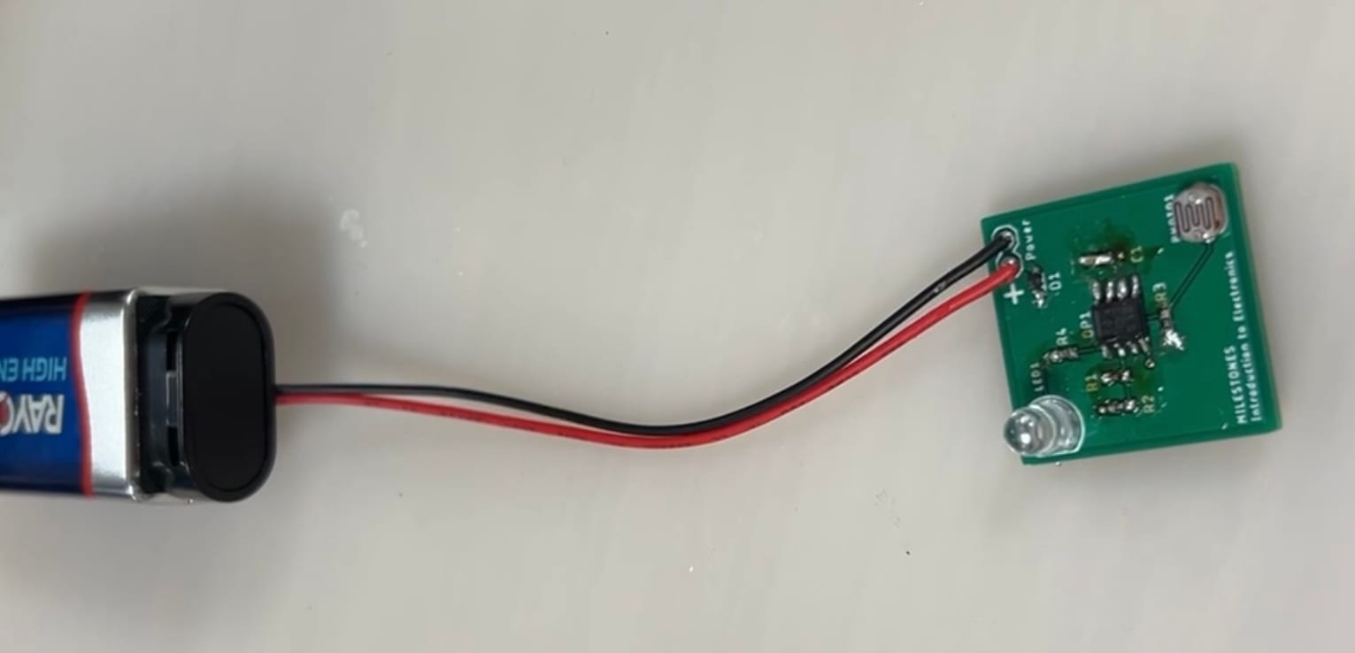 A small green printed circuit board, hand-soldered with surface-mount resistors, an 8-pin op-amp, a photocell, and a through-hole LED, powered by a 9-volt battery through red and black leads. The silkscreen reads 'MILESTONES Introduction to Electronics'.